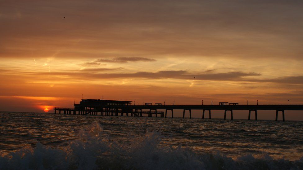 An orange and brown sky overlooks a dark pier out at sea. The sun is rising in the distance.