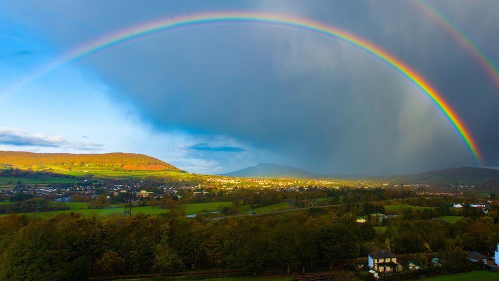 A view of rolling green countryside hills under bright blue sky and patchy clouds. A large rainbow arches over the scene, the rainbow including colours of purple, blue, green, yellow, orange and red.