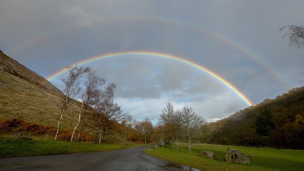 A look down a road, with steep green hills either side. Double rainbow stretch across the width of the road, with colours of purple, green, yellow, orange, blue and red.