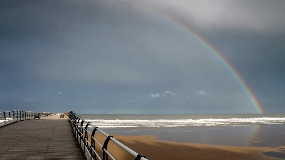 Looking down an empty pier, over a sandy beach. The sky is light blue and a rainbow stretches from the right and disappears into the horizon. Colours of the rainbow include purple, green, yellow, orange and red.