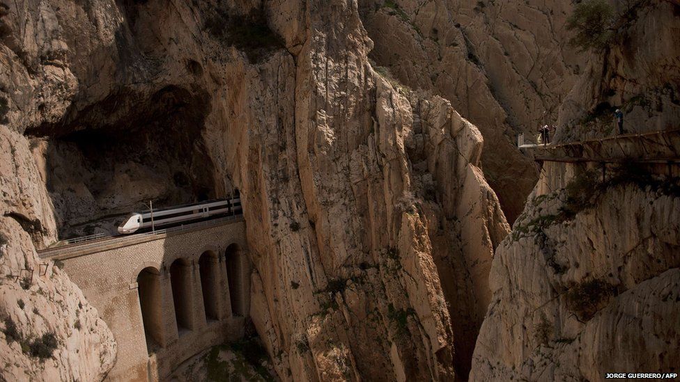 A train passes through a tunnel in the rocks as people walk on the El Caminito del Rey