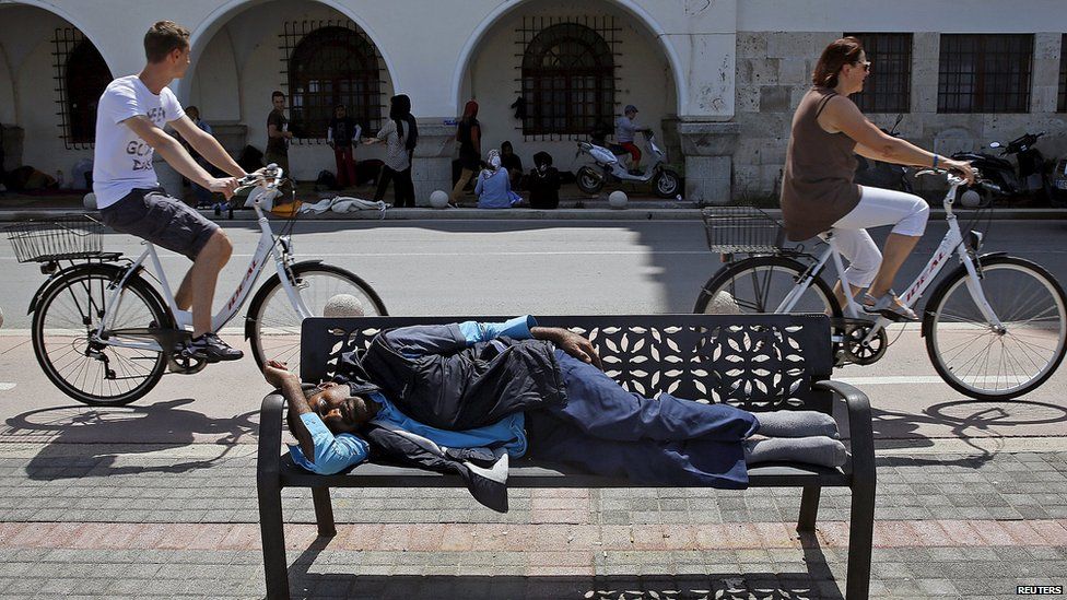 A migrant sleeps on a bench and others gather on the side of the road as two people cycle passed