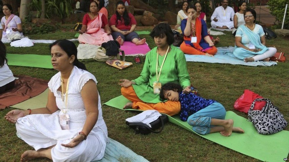 A child sleeps in a woman's lap as she performs exercises with others to participate in a prelude to the International Yoga Day at the Art of Living headquarters on the outskirts of Bangalore, India, Saturday, June 13, 2015.
