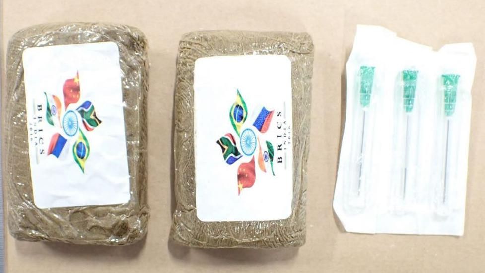 Two packages of brown-coloured drugs, wrapped in plastic and labelled as Brics India products, and three needles used on the end of syringes