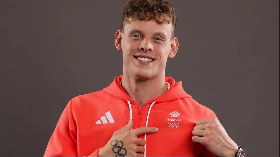 Matt Richards points at the Team GB logo on his orange Team GB hoodie while smiling at the camera.