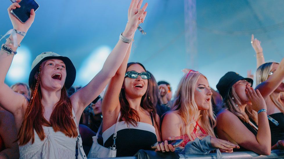 The front row of an outdoor gig, with five young women, all with long hair, raising their arms, under a blue sky