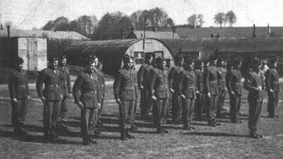 Black and white picture of soldiers standing for inspection in front of nissen huts