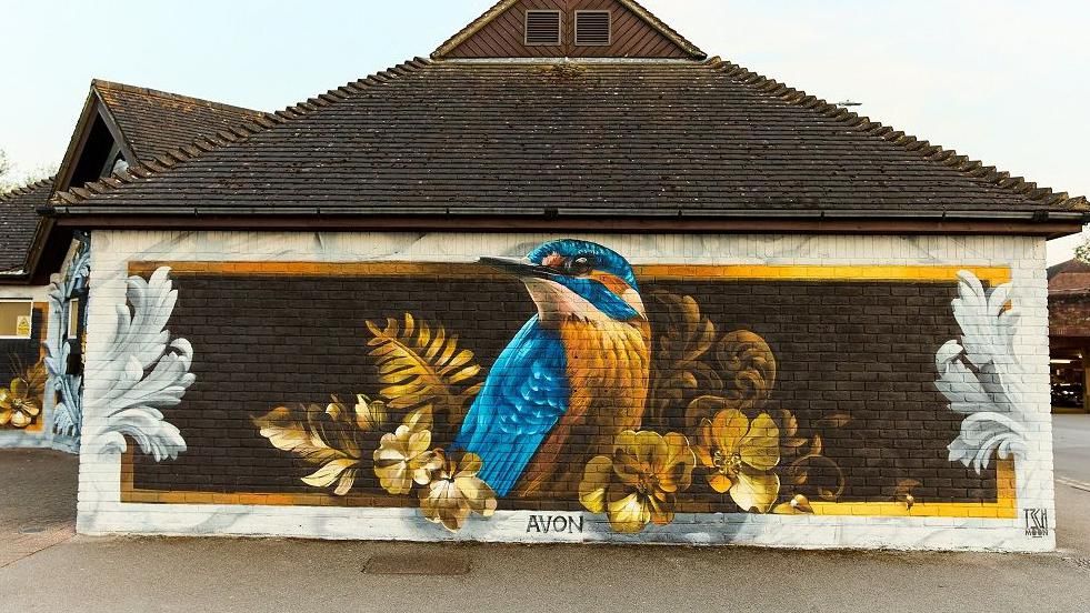 Side of a small brick building with the painted mural of a kingfisher surrounded by foliage