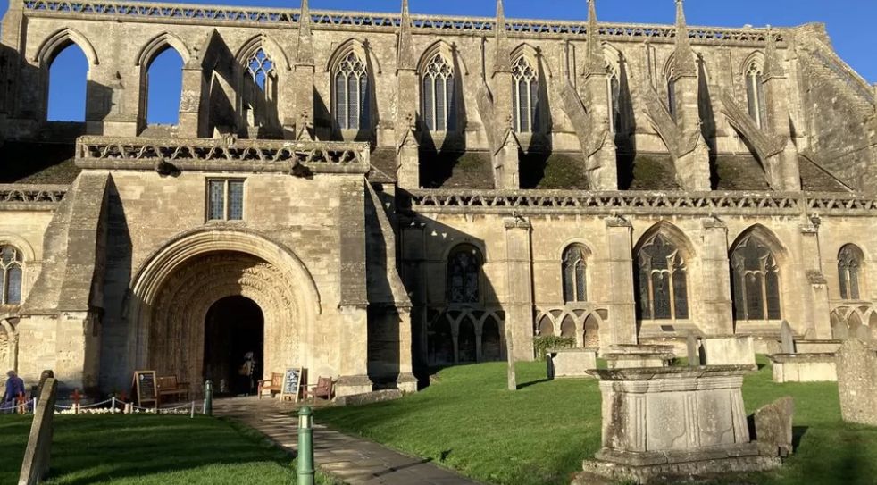 A side-on view of Malmesbury Abbey with graveyard in the foreground and a path leading to the entrance.