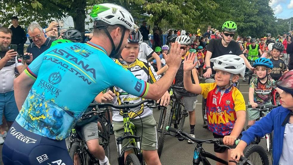 Mark Cavendish giving a child on a bicycle a high-five. Lots of other cyclists can be seen in the background.