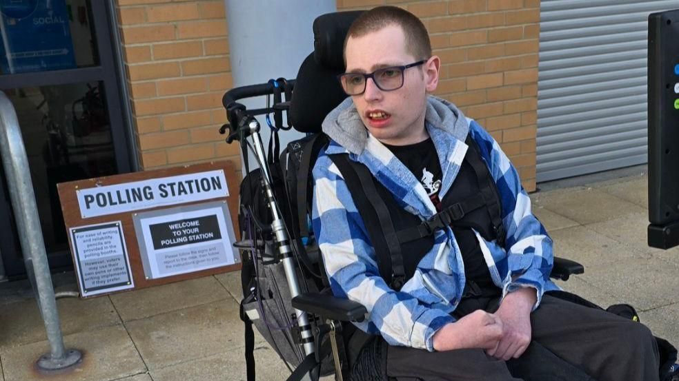 James Walker outside a polling station in his support chair wearing a blue chequered jumper, black trousers and glasses looking away from the camera