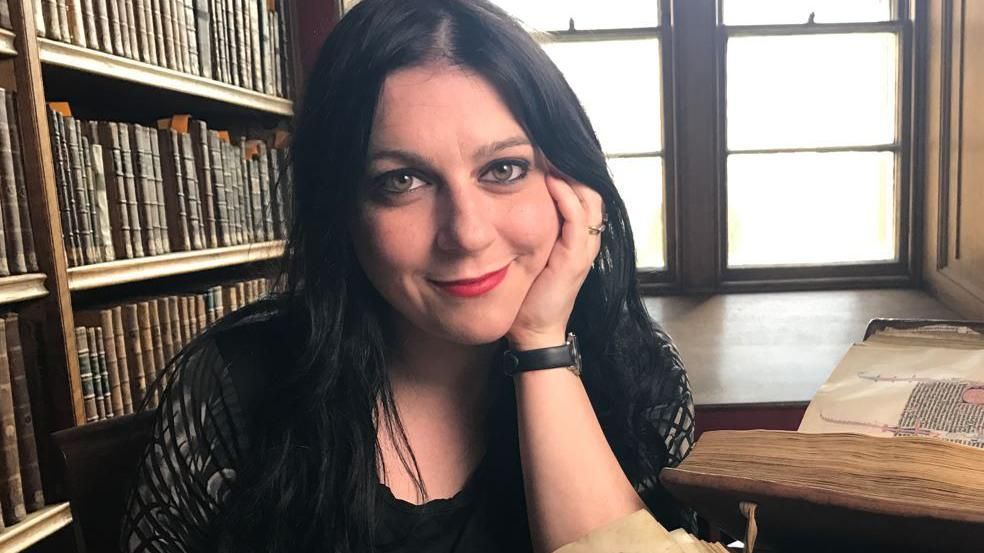 Dr Janina Ramirez sitting at a table in a library resting her head on her hand and smiling. She is wearing all black, has black hair and red lipstick