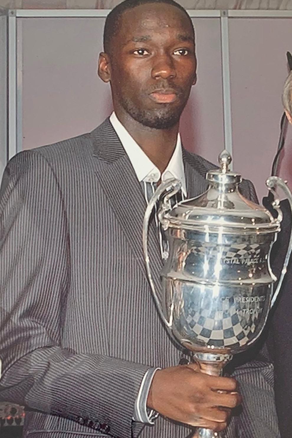 A teenage Moses Swaibu poses with a trophy while wearing a pinstripe suit