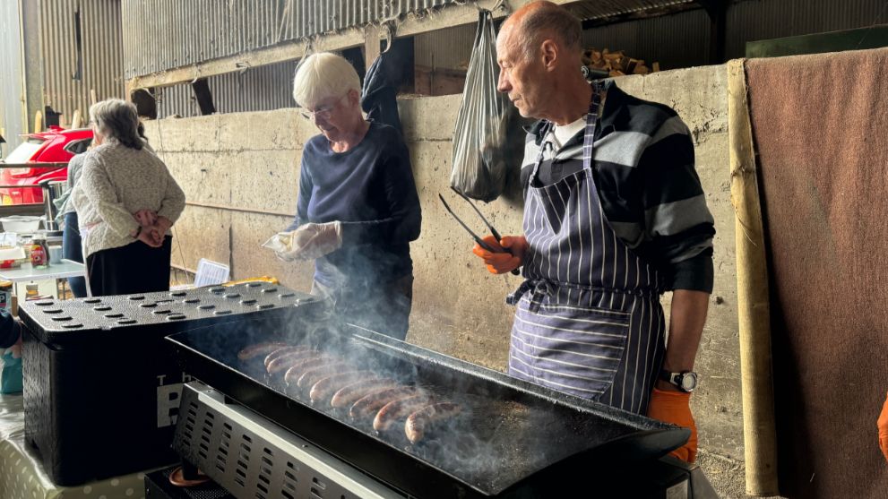 A man and a woman standing over a grill with sausages cooking on the top