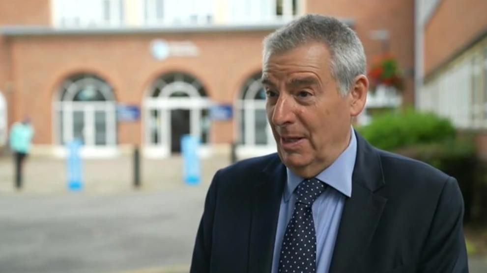 Prof David Green who has short grey/black hair and wears a blue suit over a light blue shirt with a blue tie which has white spots on it. He stands in front of a red brick building with arched windows.