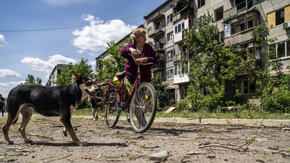 A woman leans on a bicycle near a dog, looking up at a damaged building