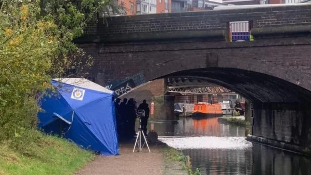 A blue police tent on the side of a canal as officers conduct searches on the towpath, under a bridge