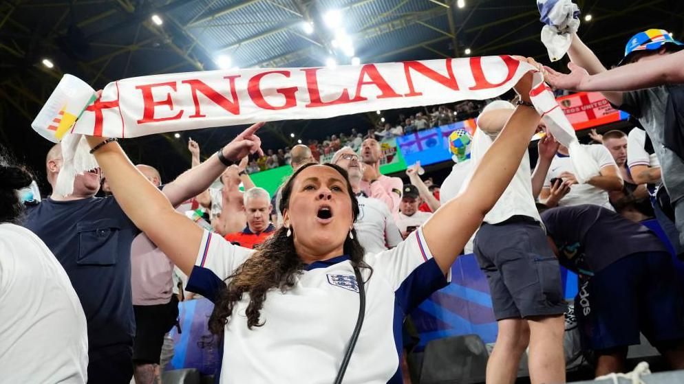 A woman in the stands of an England game holding a scarf above her head reading 'England'. She has her mouth open as if she is singing.