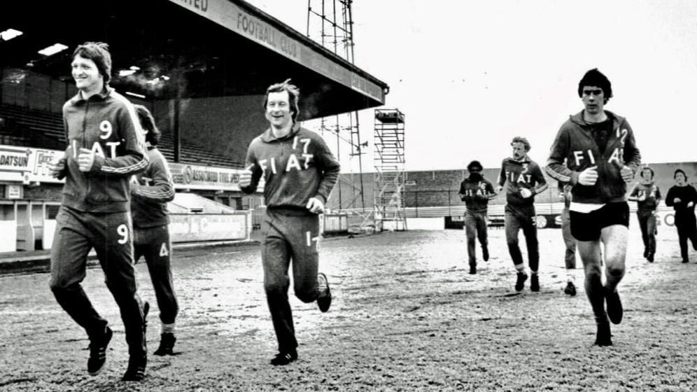 A black and white photo of nine Cambridge United players wearing tracksuits saying Fiat running at the Abbey Stadium in about the late 1970s or 1980s