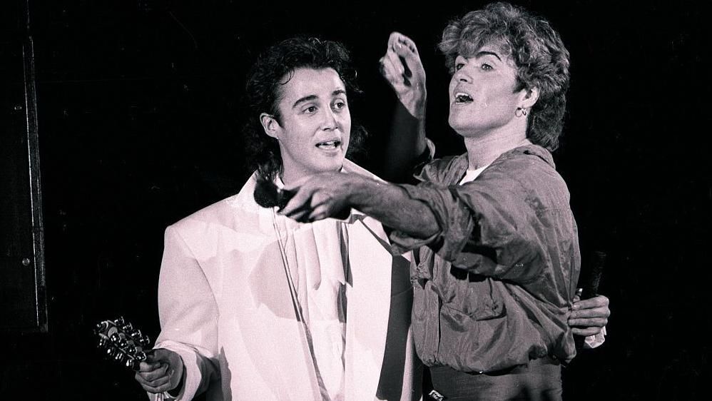 Andrew Ridgeley with slicked-back black hair and a white suit, with George Michael, who is pointing a microphone to the crowd at a concert in Whitley Bay in January 1985