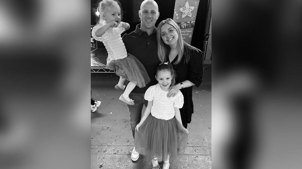 In this black and white photo, Elsie Dot Stancombe smiles at the camera, wearing a chiffon skirt and pale short sleeved top, with her dad and mum smiling standing behind her and her dad holding Elsie's younger sister Rosie. Rosie is smiling and wearing a matching outfit, holding a toy close to her mouth.