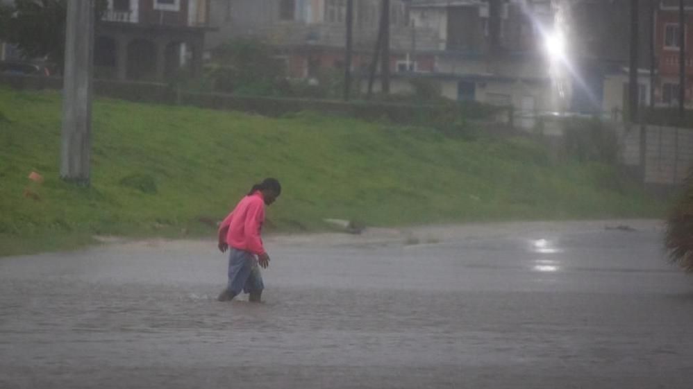 Man in bright pink jumper and shorts wades through flooded street