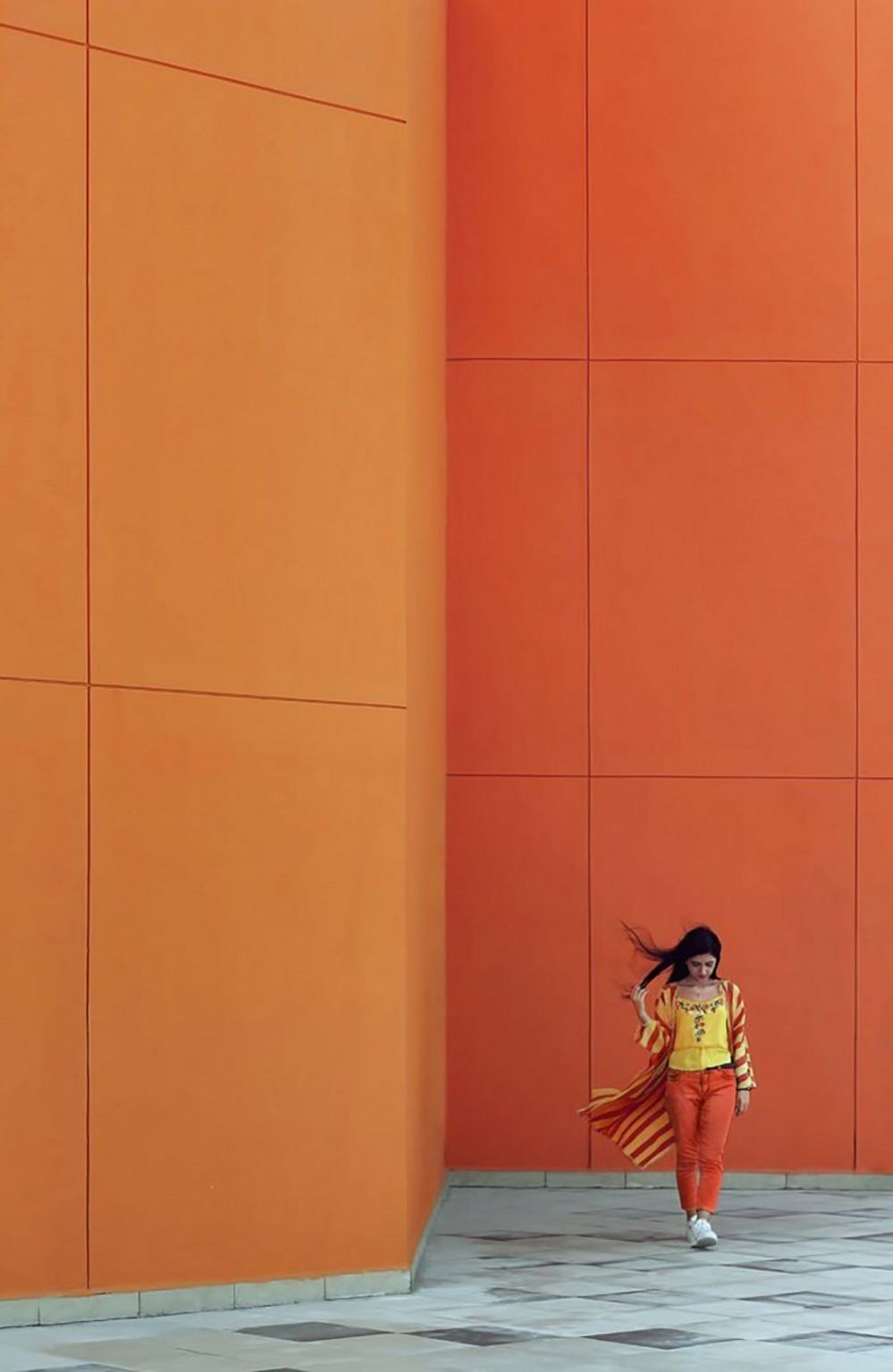 A young woman in bright orange and yellow clothing posing beside a similarly orange wall