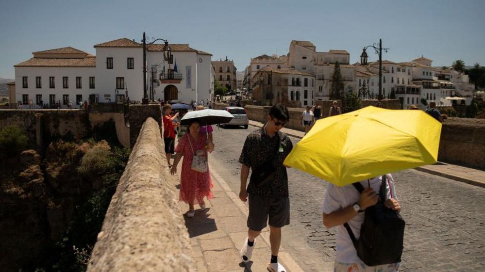 People hide from the sun with umbrellas as they cross an old bridge. White buildings in the background