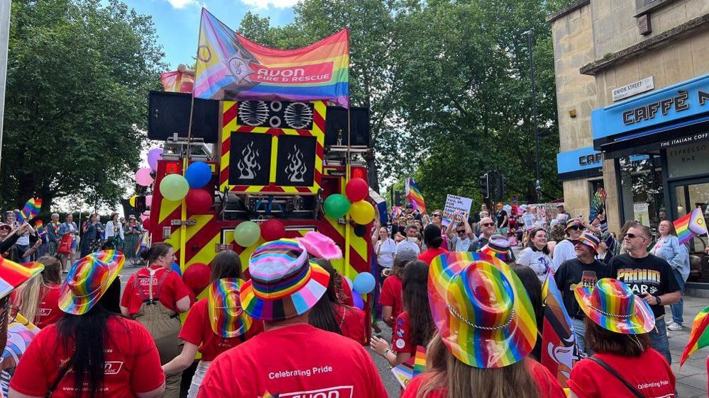 Back of a fire engine with a rainbow banner and people behind it in colourful hats