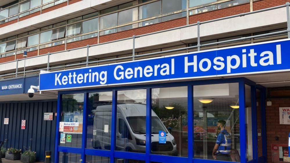 A blue and white sign that reads "Kettering General Hospital" above a glass-panelled door, with a security guard standing in a hallway. 