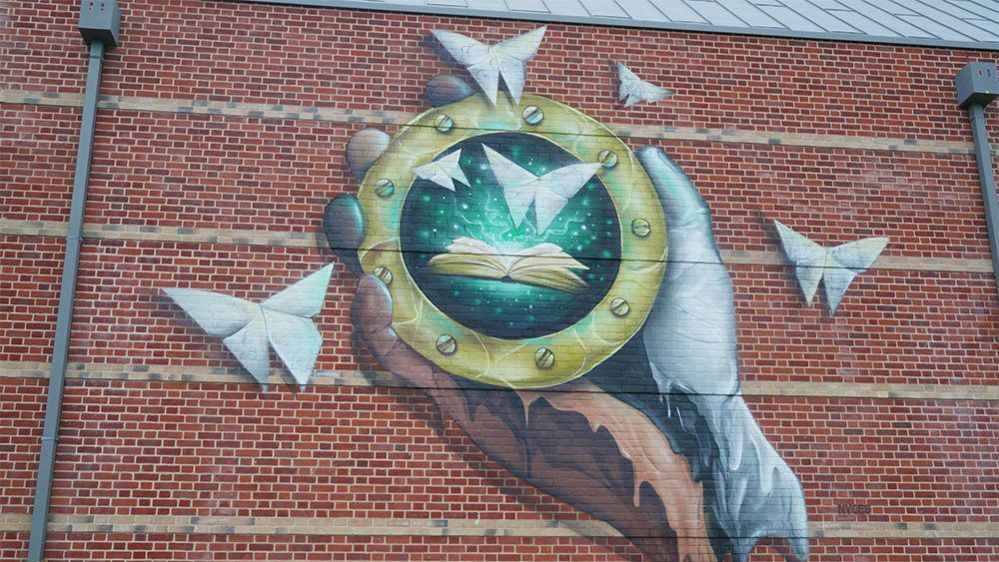 A picture shows the large scale mural created by artist Nathan Murdoch on the Suffolk Archives building The Hold. It shows a hand holding a portal suggesting the ability the time travel while origami butterflies made with folded archive records fly out of the portal.