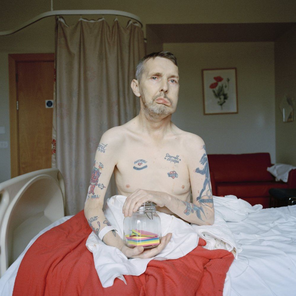 Andy, shirtless and sitting on his hospice bed holding a glass jar containing colourful paper notes
