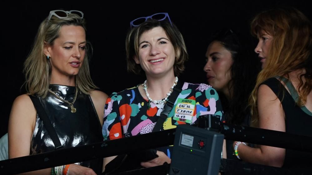 Jodie Whittaker in a crowd at Glastonbury. She is wearing a multicoloured top and is smiling