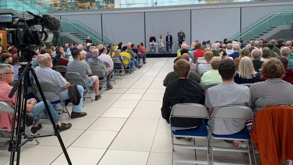 Rows of people in a hall looking at a panel of four people with one speaking into a microphone. At the back of the room is a camera on a tripod 