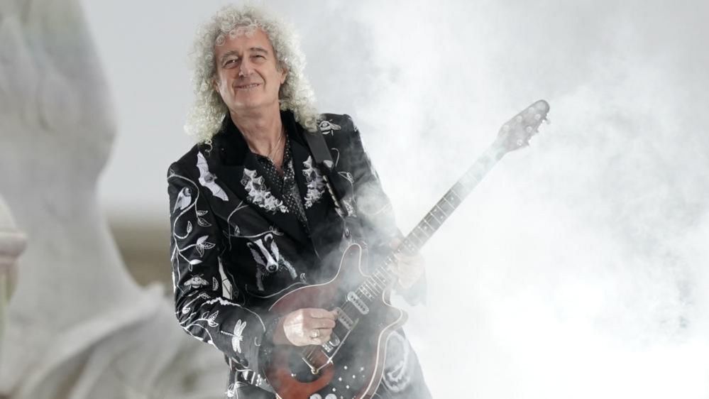 Guitarist Brian May is wearing a black and white-patterned jacket and holding an electric guitar and is surrounded by mist.