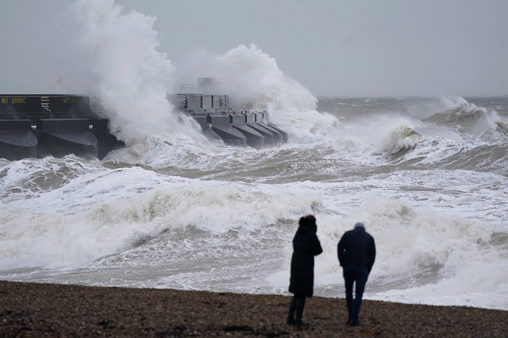 Two people silhouetted on a beach looking out to sea as strong winds and waves hit a pier on 31 December 2023