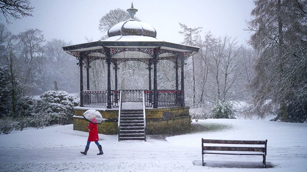 A person in a red coat holds an umbrella as they walk in snowy conditions in a park in Buxton on 8 February