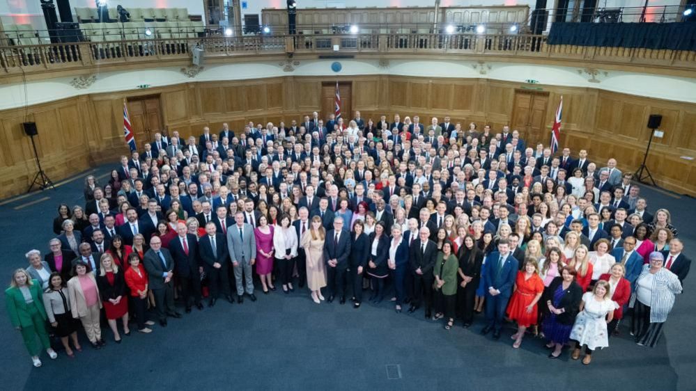 Semi-circular arrangement of new MPs around Keir Starmer in a large room with blue carpet