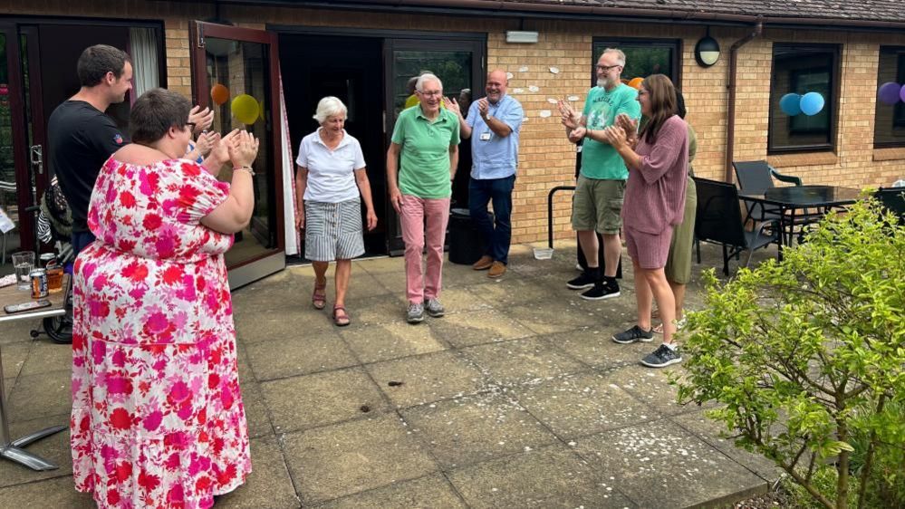 A man and a woman walk past applauding staff and volunteers into a garden area