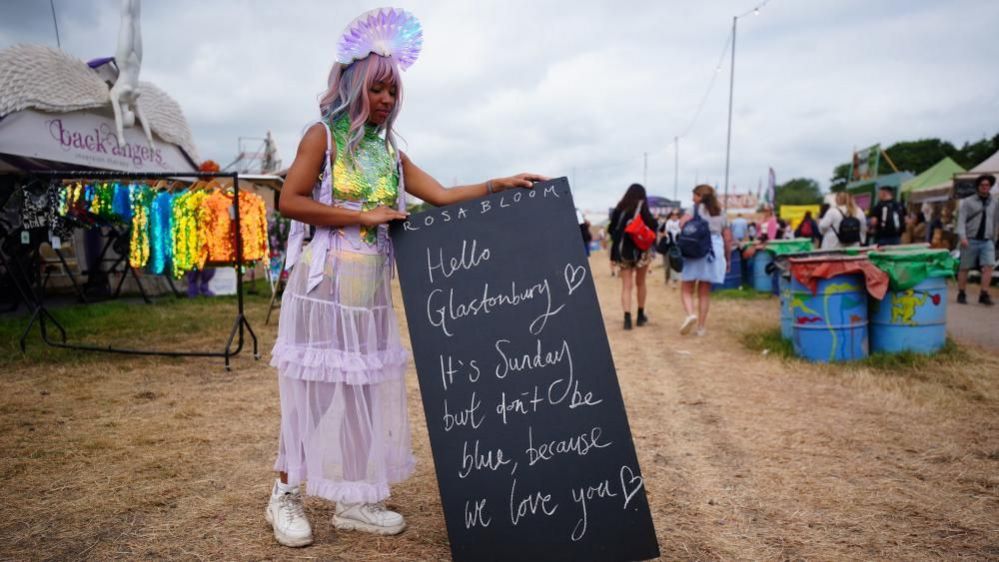 A woman dressed in purple puts out a sign reading "hello Glastonbury, it's Sunday but don't be blue because we love you"