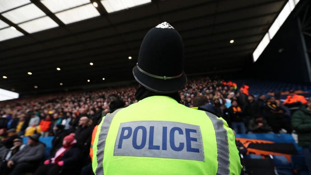 A police officer with his back to the camera looks toward a stand filled with football supporters