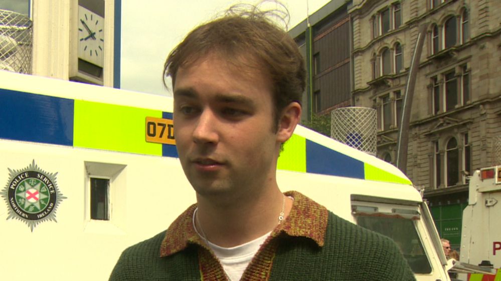 Karl is standing with a police jeep behind him. He's wearing a green jumper.