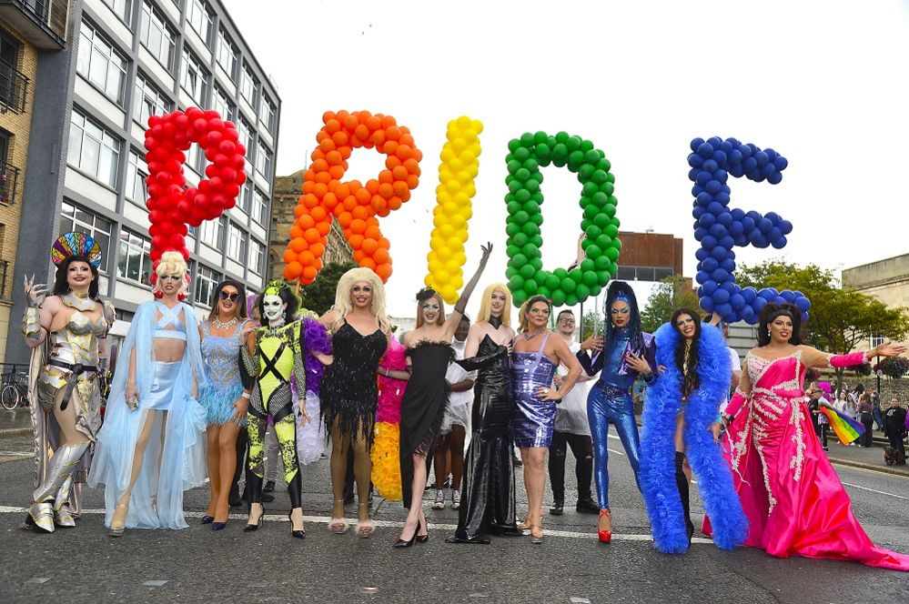 A group of drag queens stand in front of the pride sign