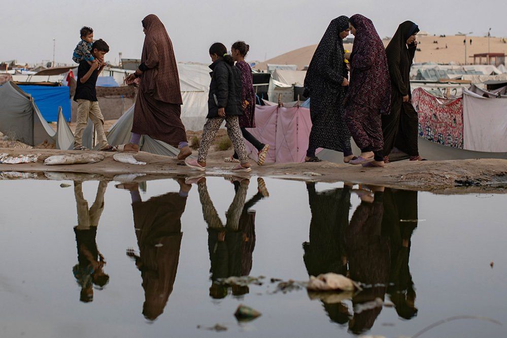 People are reflected as they walk in front of a sewage spill in a makeshift camp on the outskirts of Rafah on 26 April
