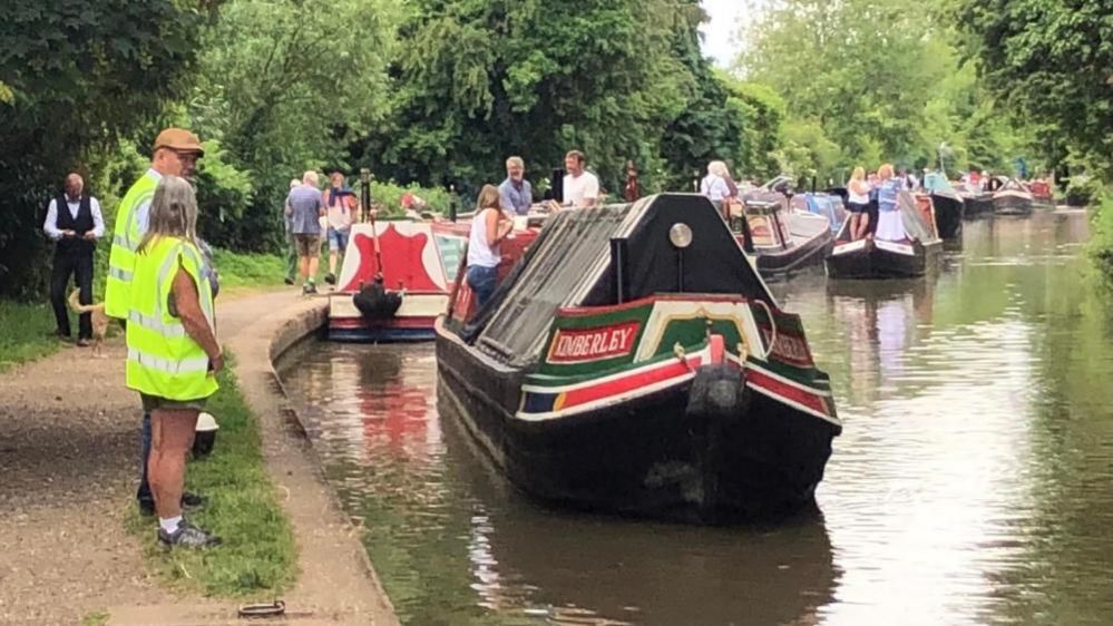 Collection of narrowboats with people aboard the boats and on the towpath