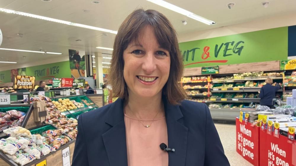 Rachel Reeves with medium-length dark hair and wearing a dark jacket and pink top standing in a supermarket