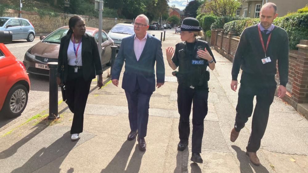 Two men and two women walk along a wide pavement in Raunds. Lee Barron is wearing a blue suit, while Insp Kiernan is wearing a police uniform. They are casting long shadows on the pavement.