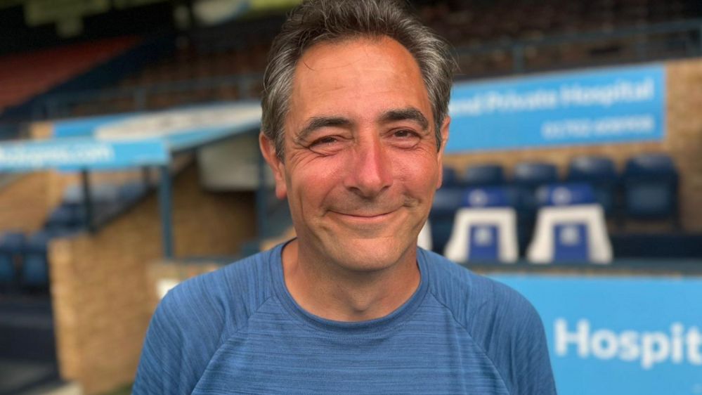 Karl Lansley, wearing a blue top and looking tanned after his marathon walk, smiles as he stands in front of one of the stands at Southend United's ground