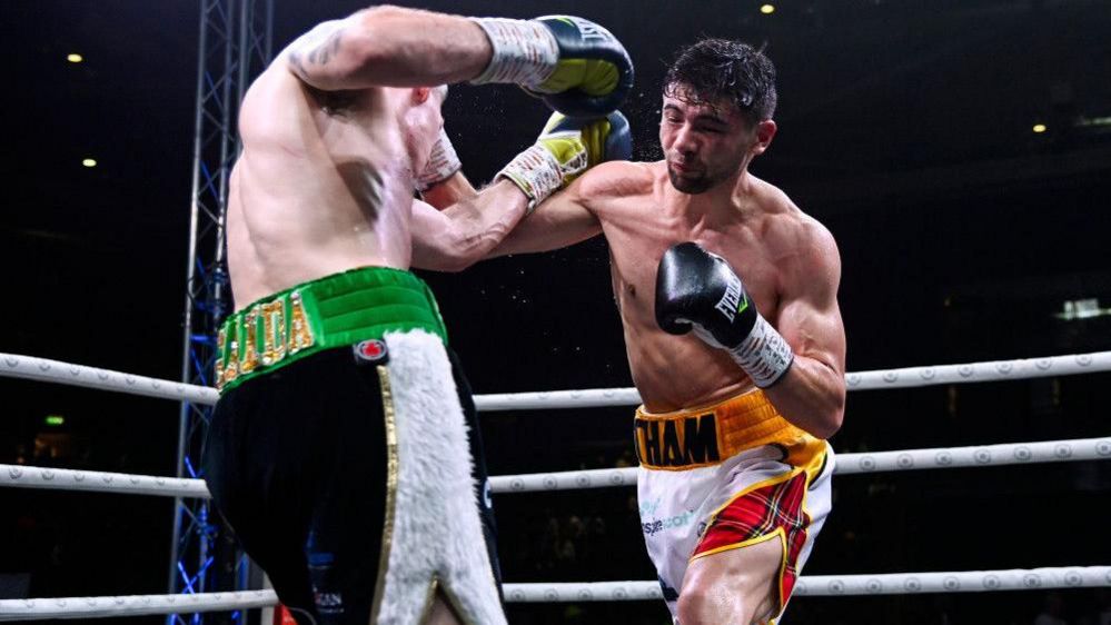 Andy Tham competing in a boxing ring, wearing white shorts with tartan detailing, punching another boxer wearing black and white shorts with green trim.