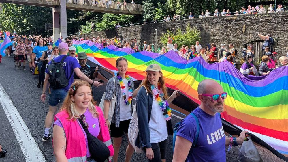 Crowd carries a long piece of rainbow material through a street